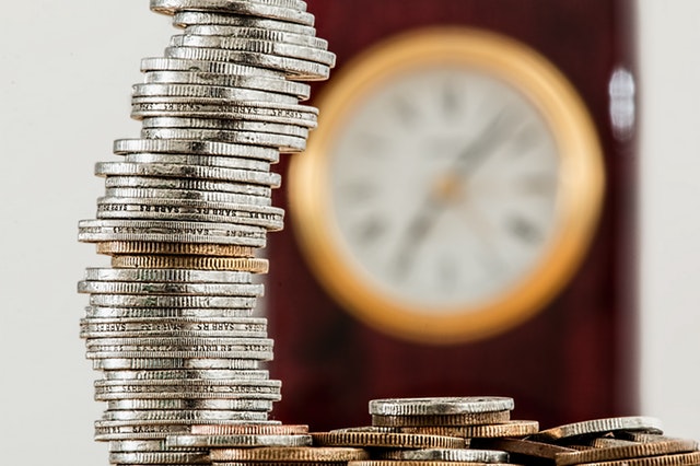 A stack of coins sits in the forefront, and in the background is a blurred clock hanging on a wall.