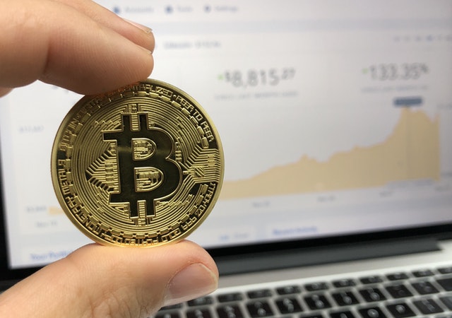 A golden-colored coin with a large "B" in the center, held up between two fingers in front of a computer screen with graphs and other figures.