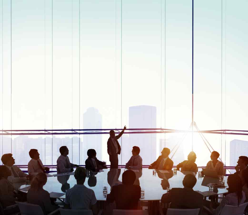 Silhouettes at round table look out large window where man standing points.